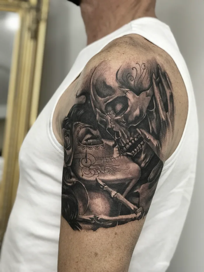 Black and grey skull kissing woman upper arm tattoo by Esther, Unicorn Tattoo Art Collective, Ashford Surrey