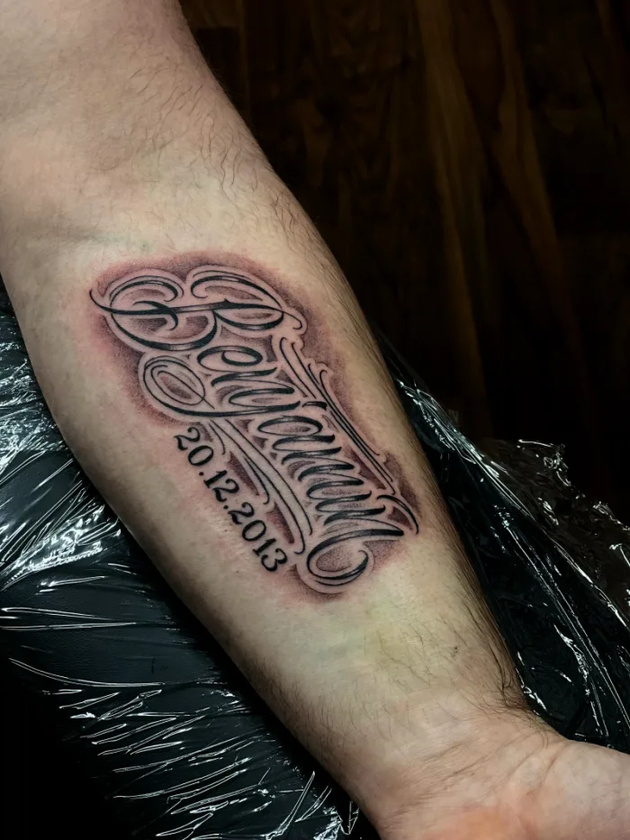 Custom black and grey name and date lettering forearm tattoo in chicano script style by Esther, Unicorn Tattoo Art Collective, Ashford Surrey