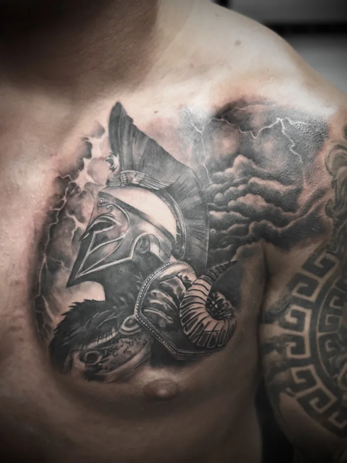 Black and grey Spartan warrior chest tattoo with helmet, armour, clouds and lightning by Esther at Unicorn Tattoo Art Collective in Ashford Surrey