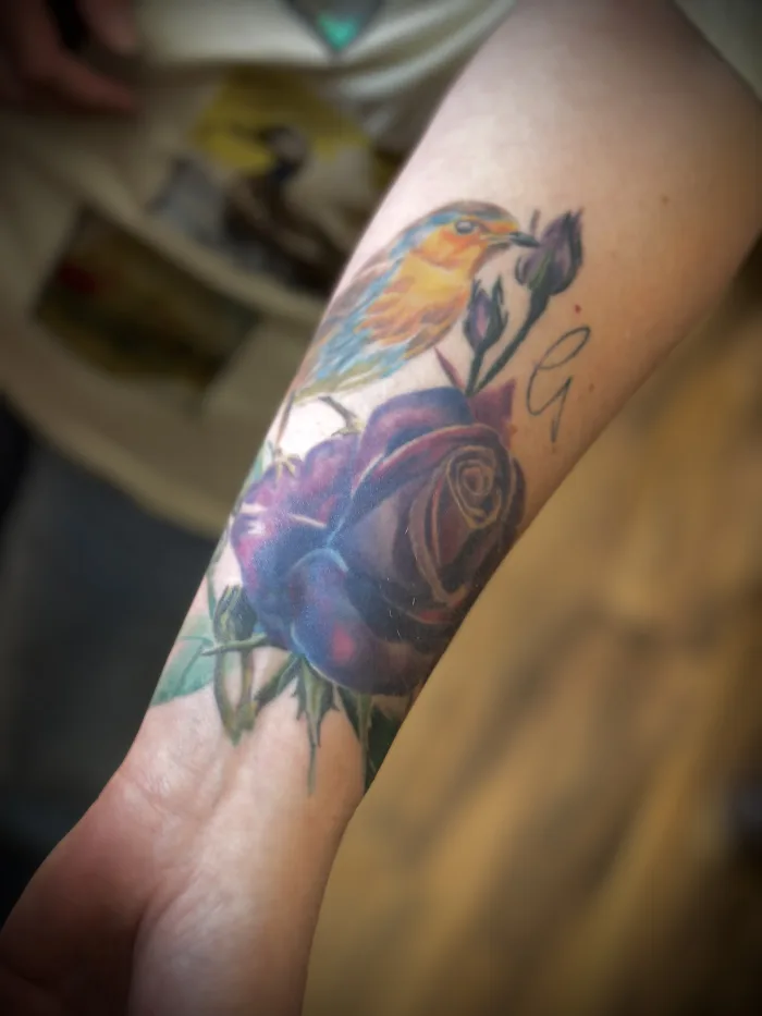 Healed colour realism bird and rose tattoo by Esther at Unicorn Tattoo Art Collective in Ashford, Surrey, featuring a colourful bird, purple rose and floral details on the forearm