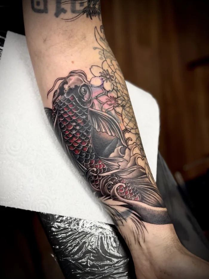 Second-session progress photo of a forearm cover-up by Esther, featuring a koi fish with red accents, Japanese waves and cherry blossoms.