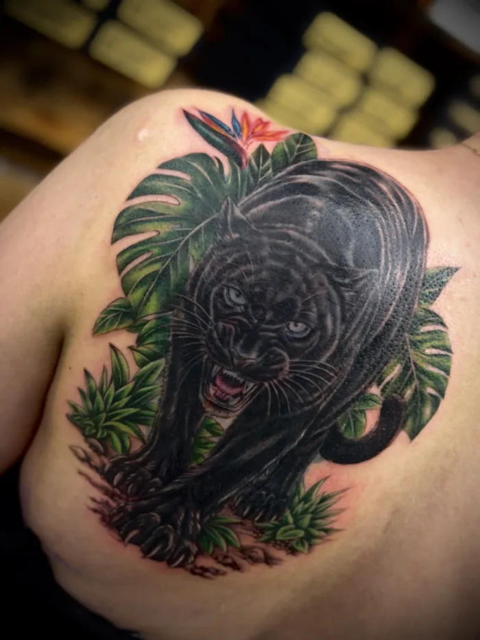 Black panther cover-up tattoo on the upper back by Esther, featuring green tropical leaves and a colourful bird of paradise flower.